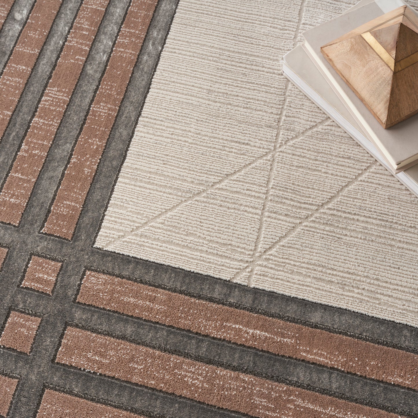 Beige geometric area rug with brown and charcoal grid patterns; stacked books and a wooden pyramid ornament in corner.