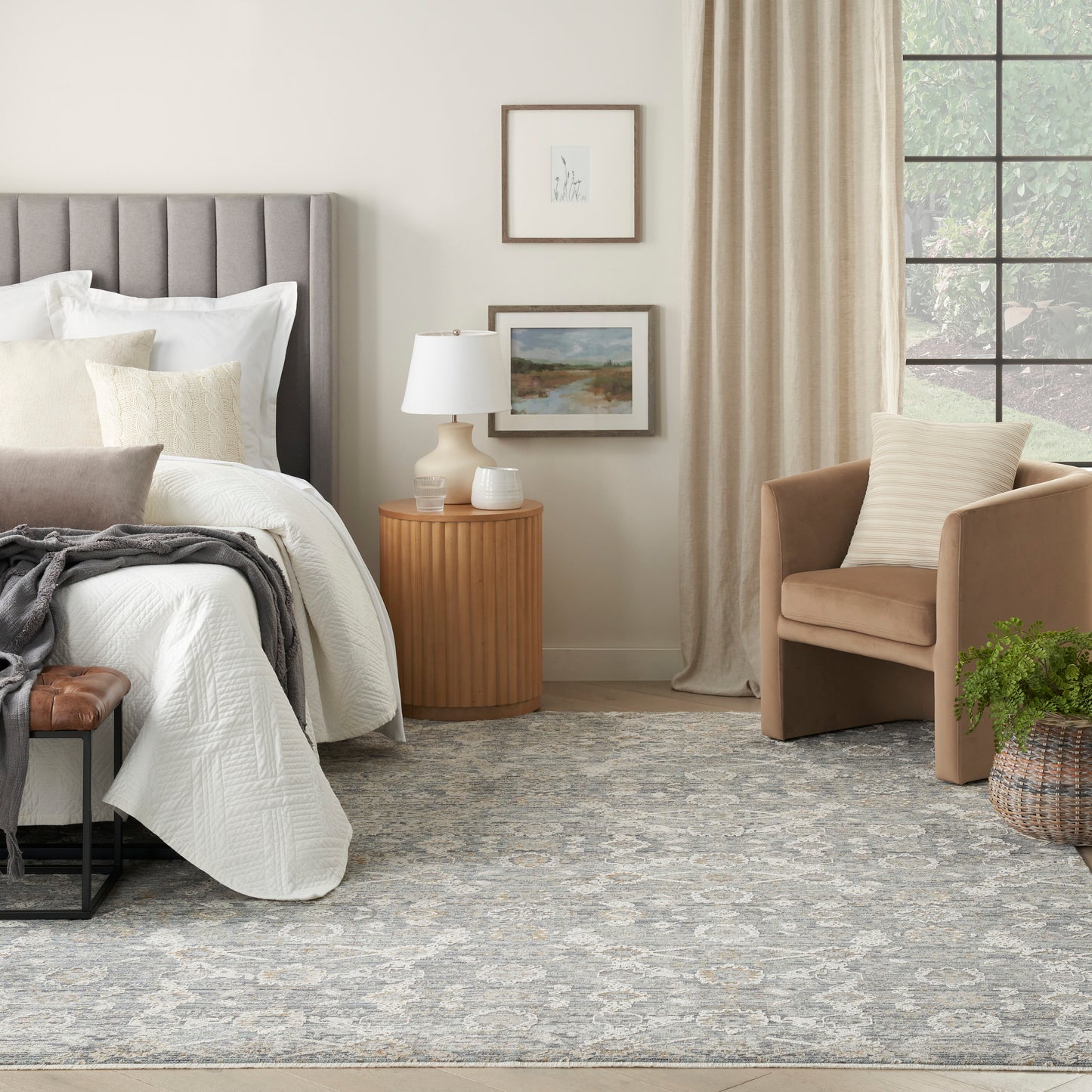 Neutral bedroom with gray tufted headboard, white quilted bedding, beige armchair, round wood nightstand, framed art, blue-gray rug (5' x 8').
