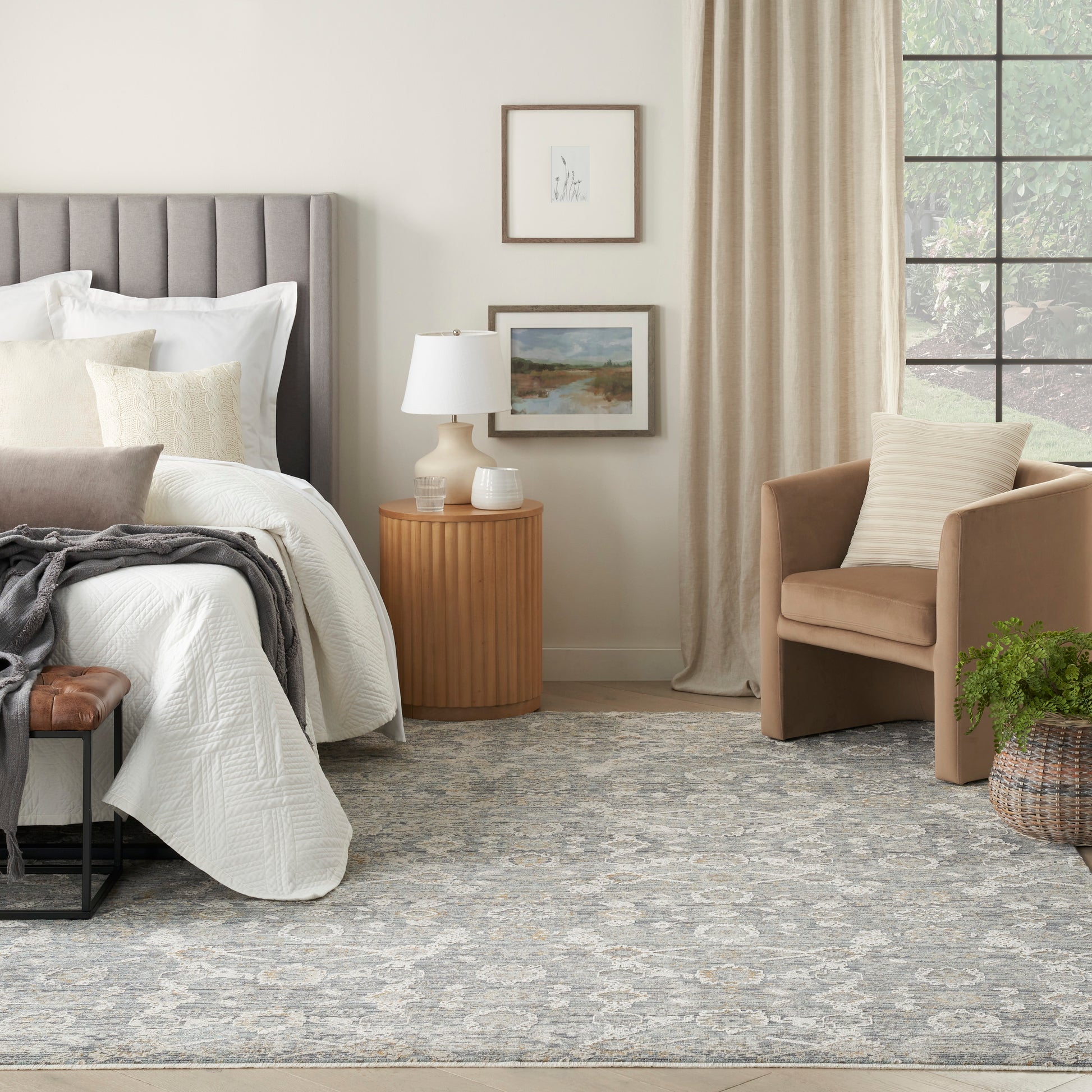 Neutral bedroom with gray tufted headboard, white quilted bedding, beige armchair, round wood nightstand, framed art, blue-gray rug (5' x 8').