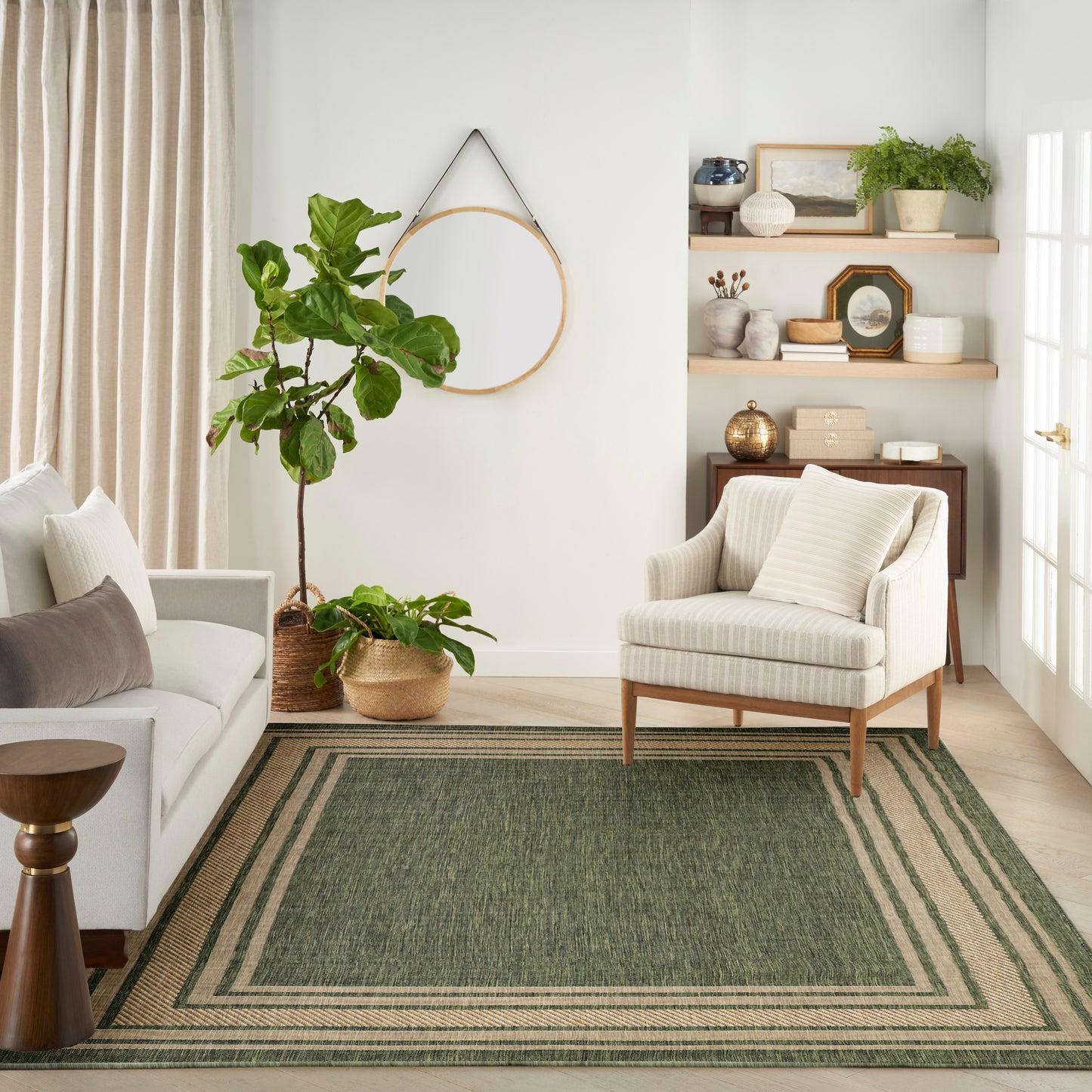 Bright cozy living room with cream sofa, beige armchair, green rug with beige border, and potted plants on wooden shelves.