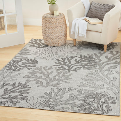 Light gray area rug with dark gray coral pattern, paired with a cream armchair, woven side table, plant, and knit throw.