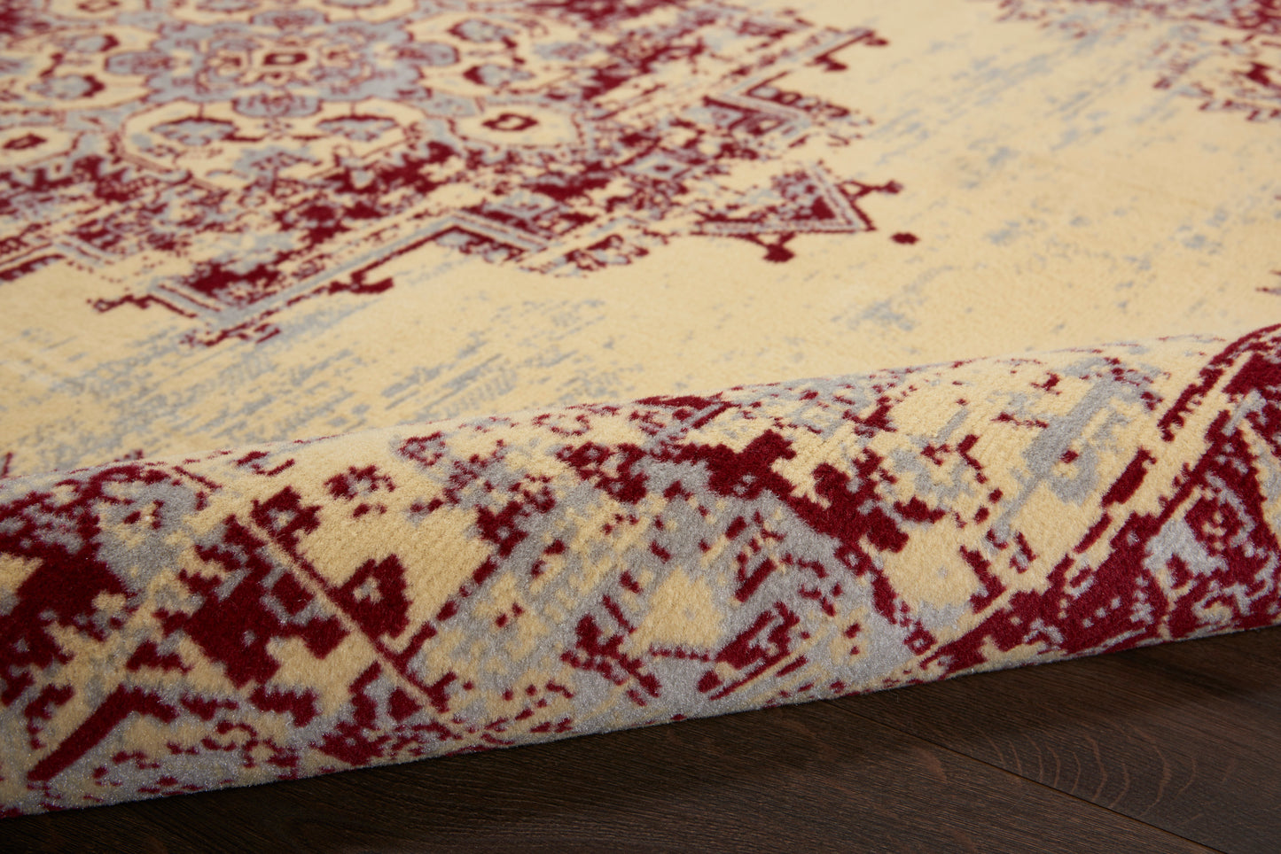 Beige oriental rug with red floral motifs and pale blue-gray accents; rolled corner shows plush pile on dark wood.