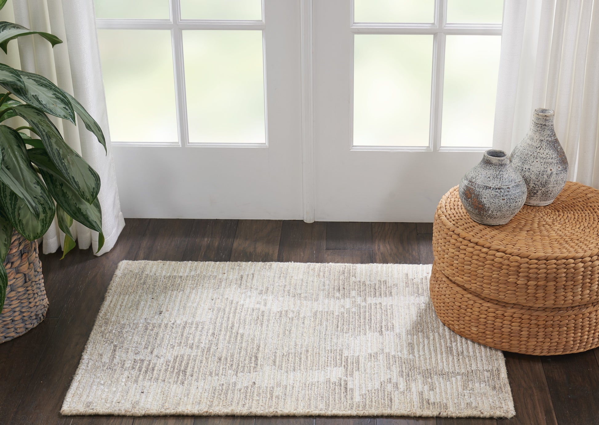 Neutral beige rug on dark wood floor beside a round woven ottoman, vases, and a potted plant by glass doors.