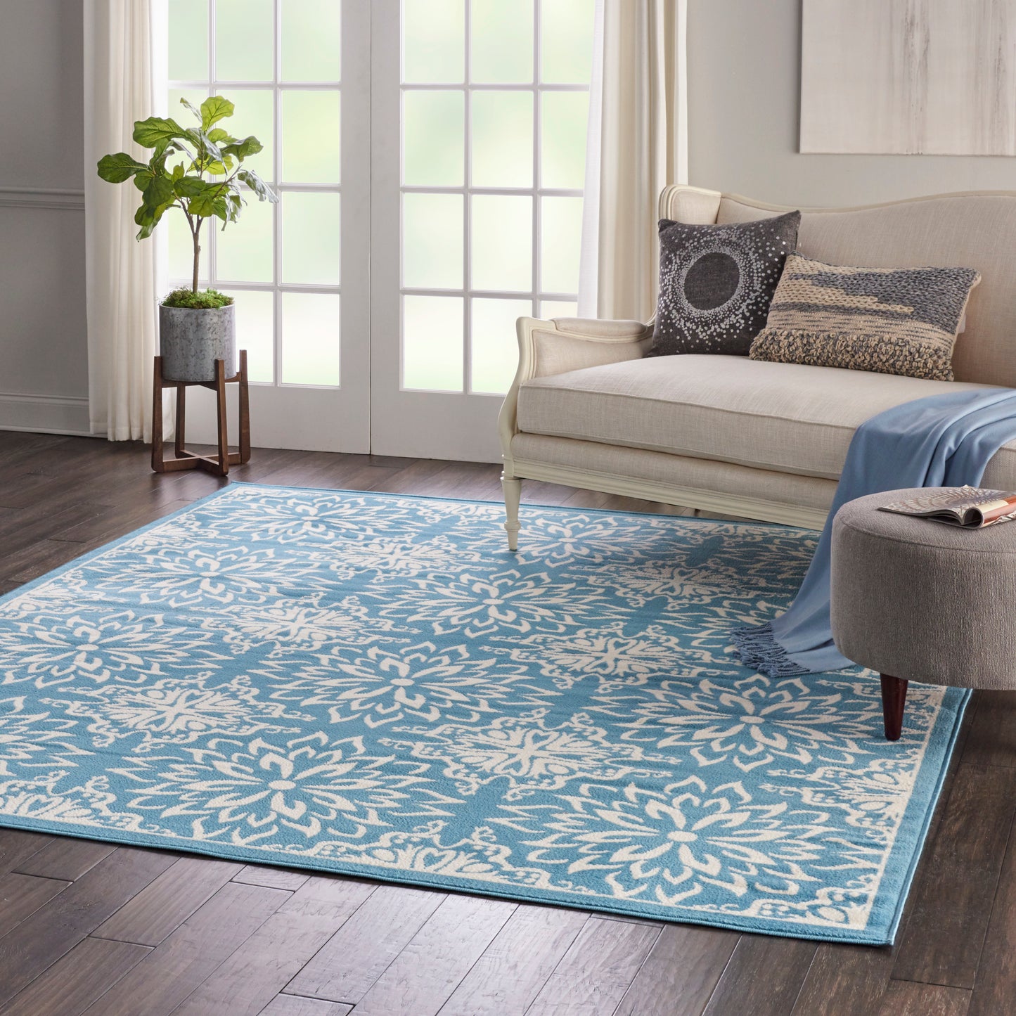 Beige sofa with blue throw and printed pillows; blue floral rug, ottoman, and potted plant near French doors.