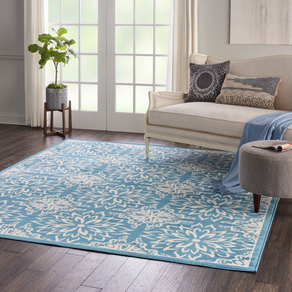 Beige sofa with blue throw and printed pillows; blue floral rug, ottoman, and potted plant near French doors.