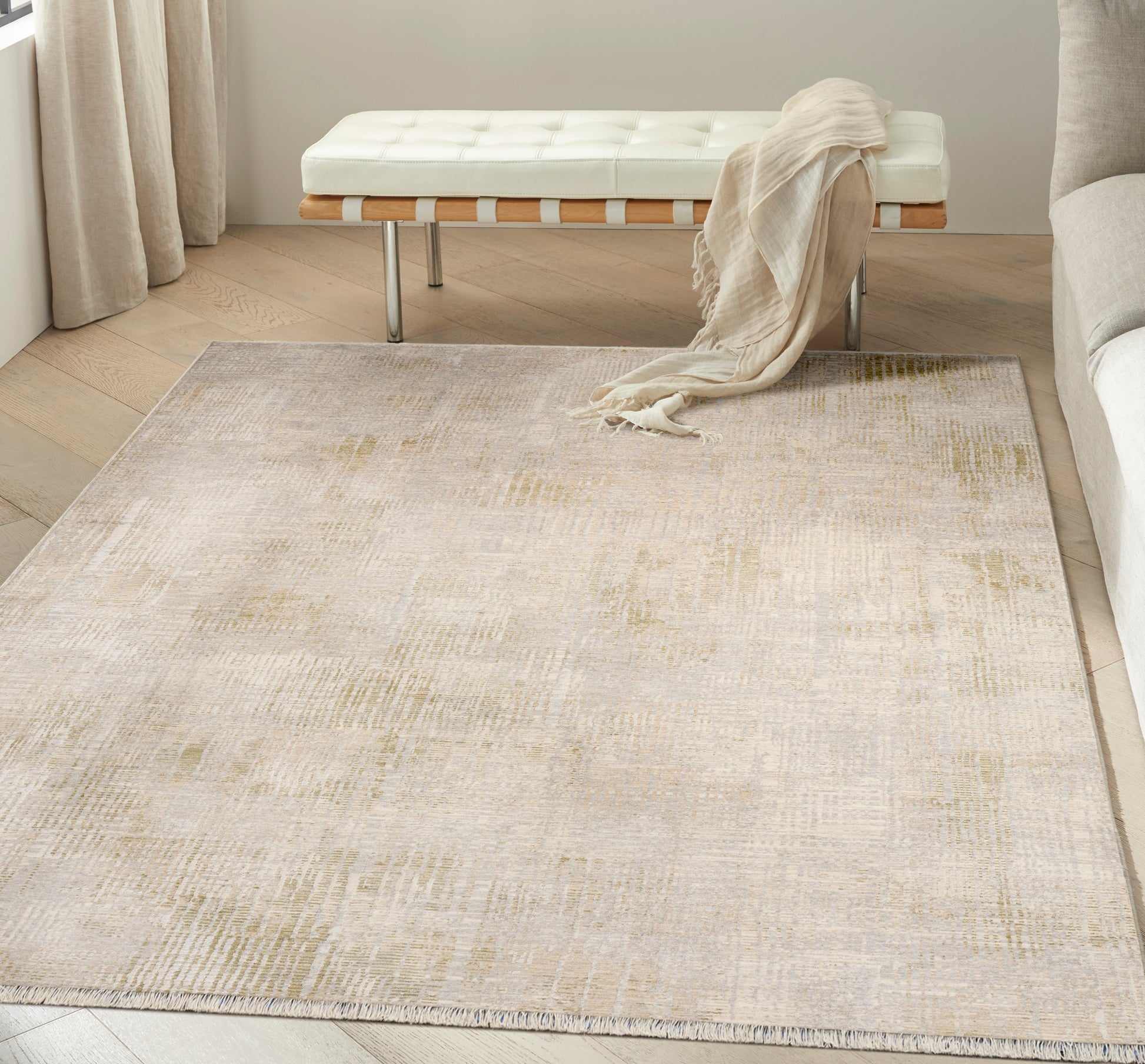 Neutral beige area rug with subtle abstract pattern on light wood floor; white tufted bench and beige throw.