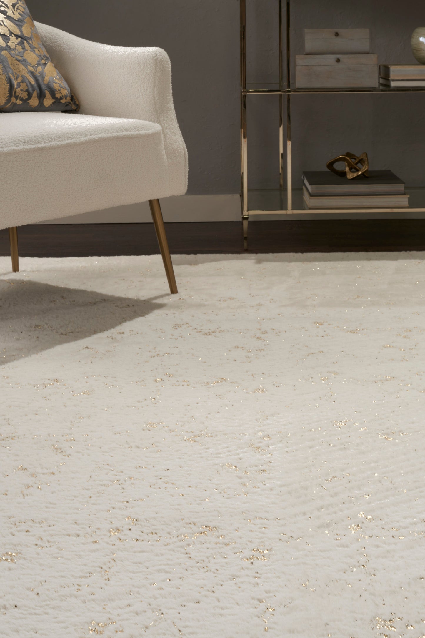 Cream boucle armchair with wooden legs beside a glass-and-brass shelf; ivory textured rug with subtle gold flecks.