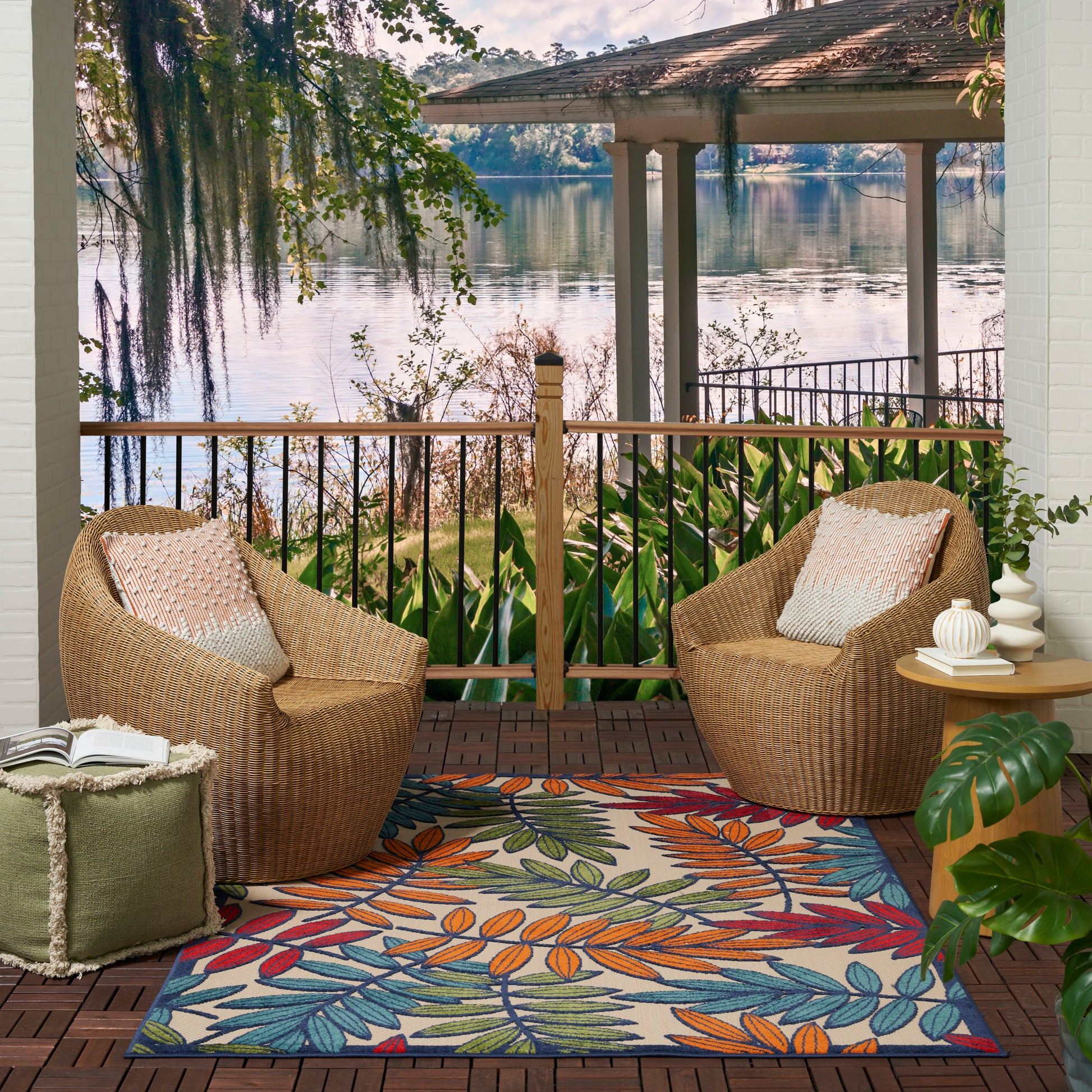 Outdoor tropical leaf rug (5' x 8') on a wooden deck with a lounge setup—two rounded chairs and a side table, lake view.