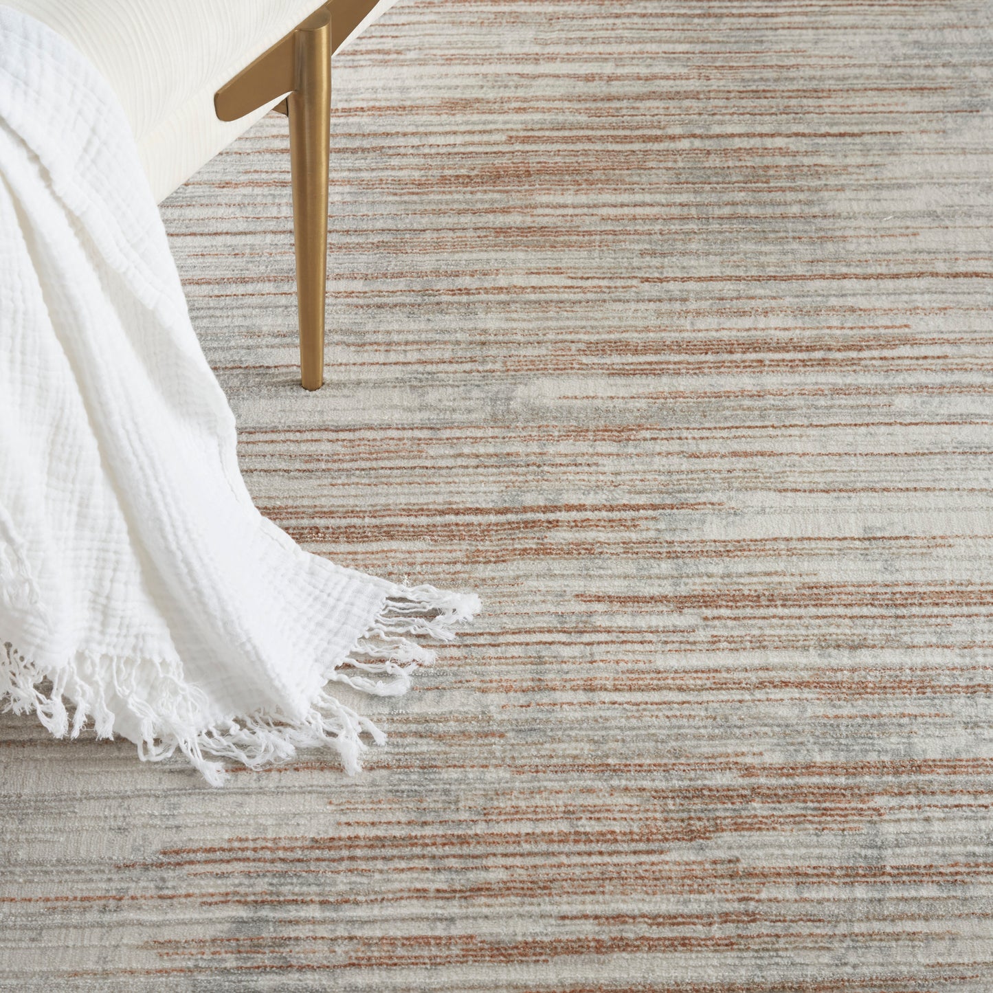 Beige striped area rug with orange-brown lines; white fringed throw blanket draped over a gold metal furniture leg.