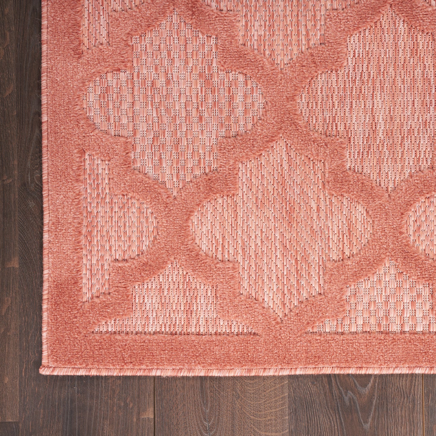 Coral-pink woven area rug with interlocking geometric diamonds and textured border on dark wood floor.