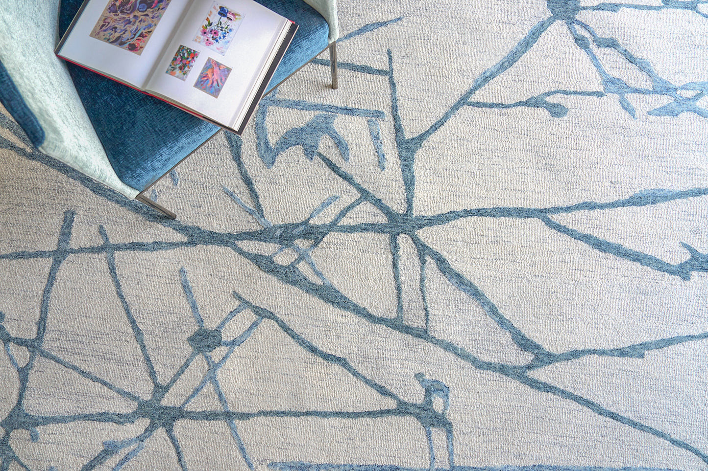 Beige rug with blue abstract branch pattern; modern style, shown with a blue velvet chair and an open art book.