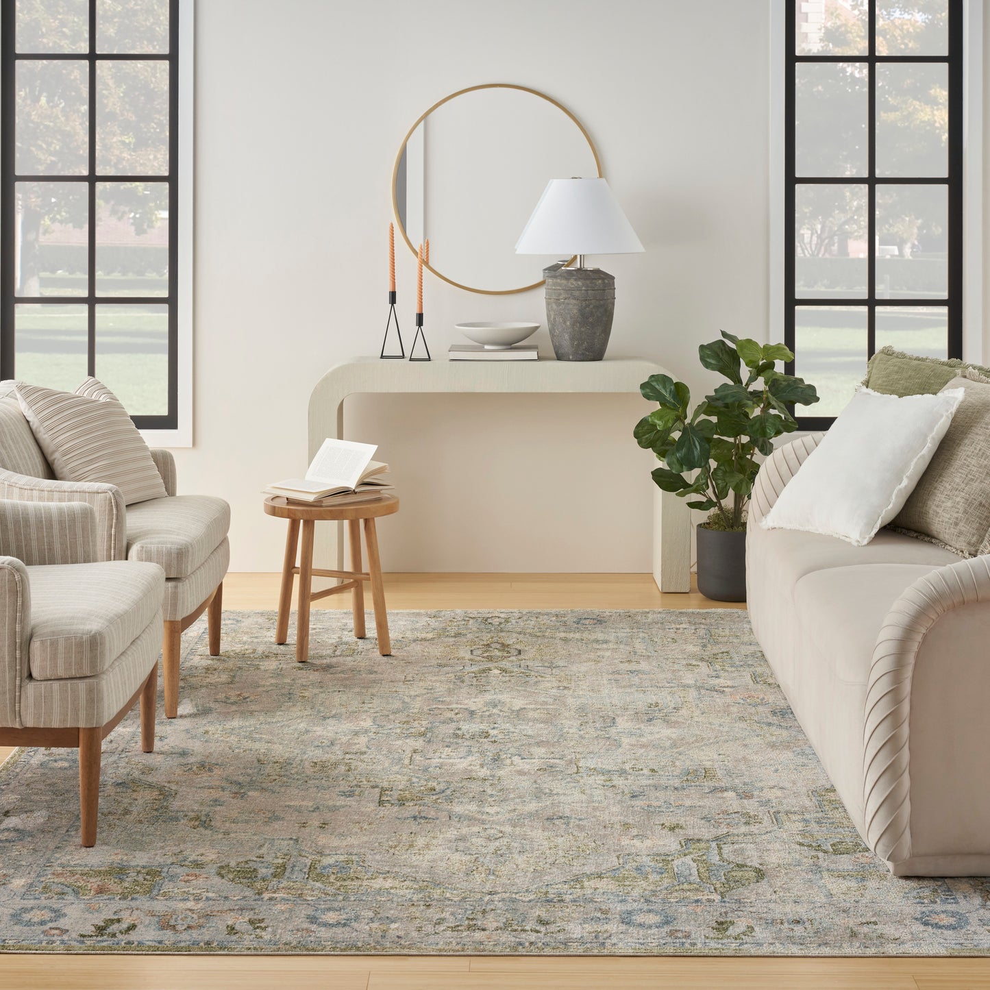 Neutral living room with beige sofa, striped armchairs, round wood stool, patterned rug, console table, gold round mirror, lamp, plant.