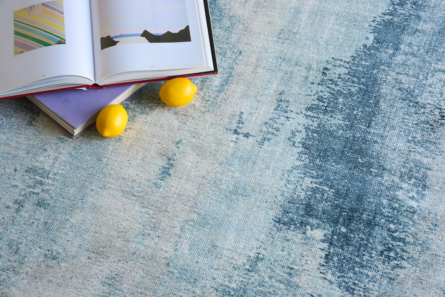 Blue textured woven area rug with abstract wash pattern; open art books and two yellow decorative balls sit on it.