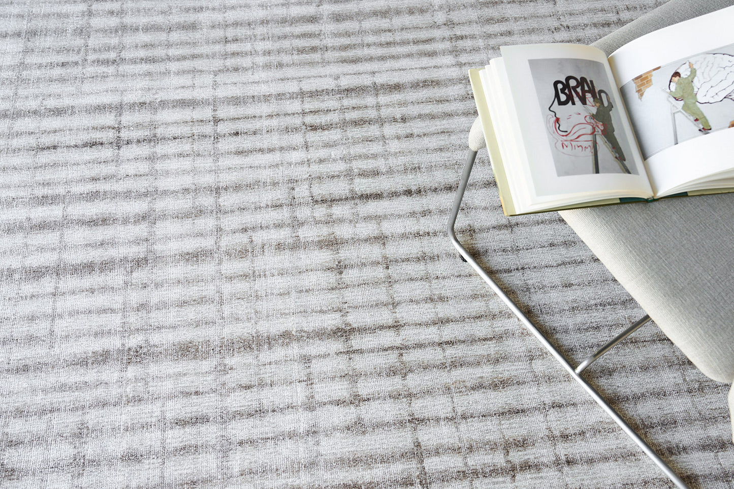 Light grey textured rug with a metal-framed chair and an open art book resting on the chair.