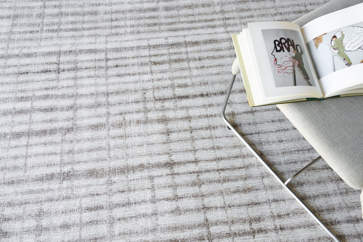 Light grey textured rug with a metal-framed chair and an open art book resting on the chair.