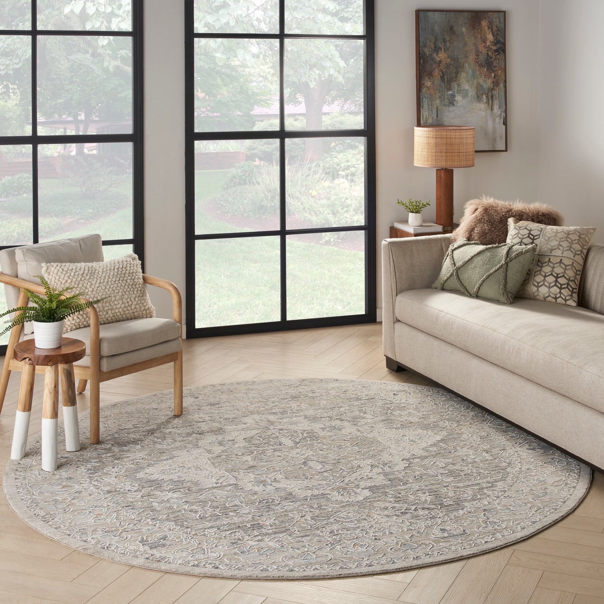Round gray-patterned rug (8' diameter) on the floor, with a beige fabric sofa, a light-wood chair, black-framed windows, and a plant on a side table.