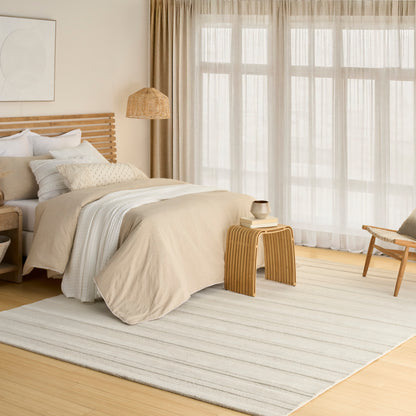 Neutral bedroom with beige bedding, wooden slatted headboard, woven pendant lamp, striped rug, sheer curtains.