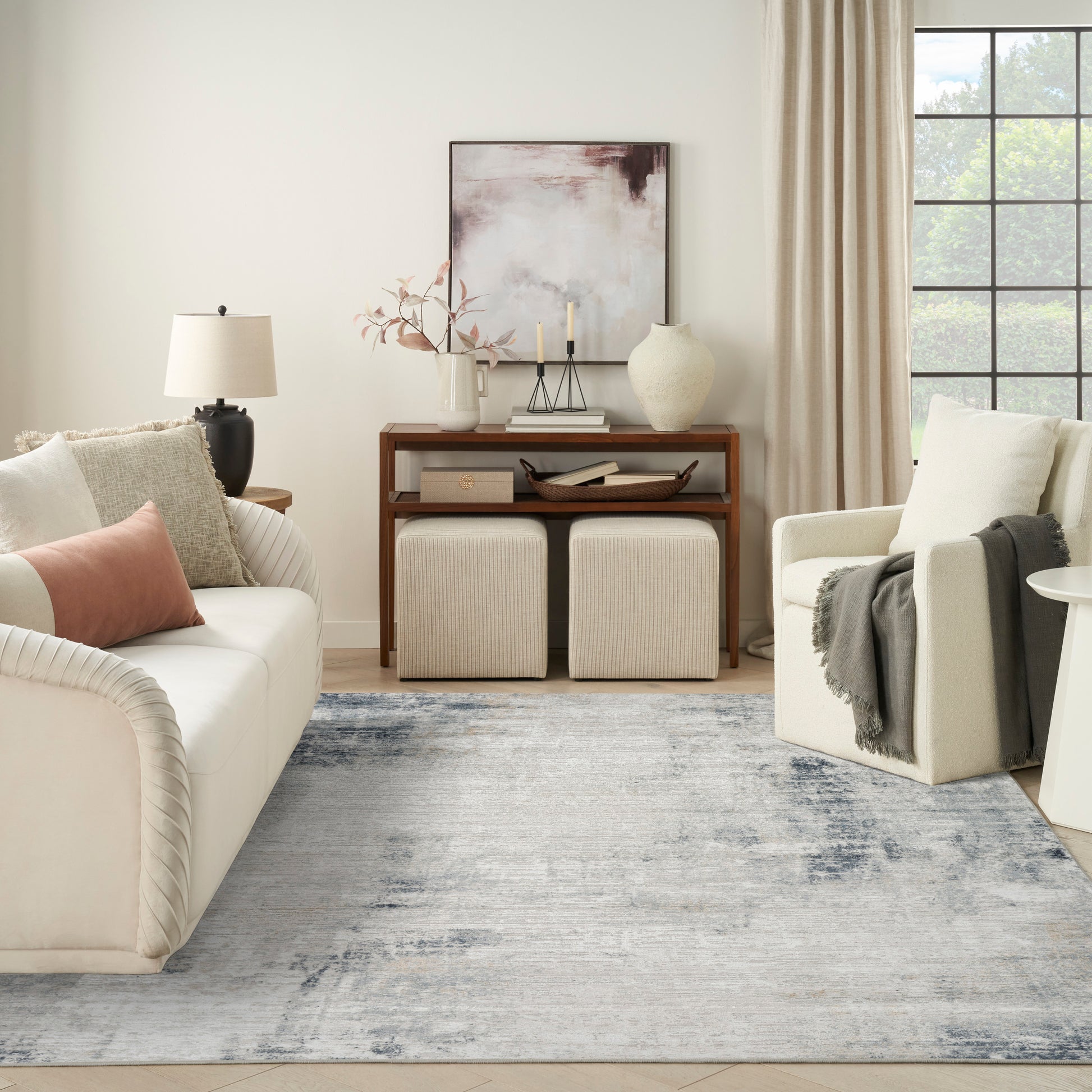 Neutral living room with cream sofa, white armchair, light wood console, two textured ottomans, blue-gray rug.