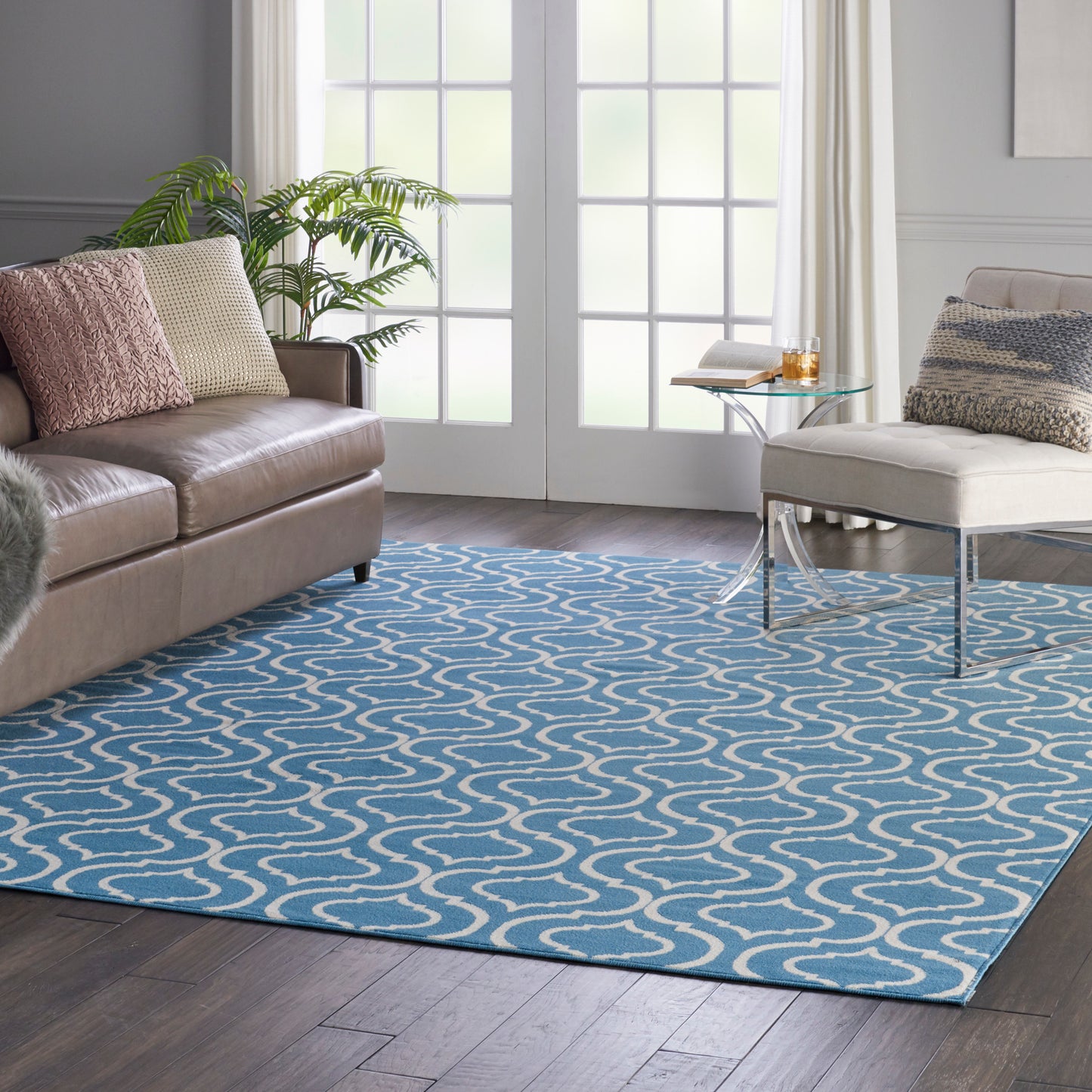 Living room with blue rug featuring white wavy lines, tan leather sofa, beige cushions, glass coffee table, and greenery.