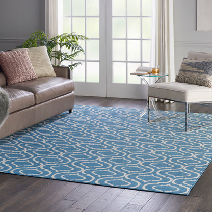 Living room with blue rug featuring white wavy lines, tan leather sofa, beige cushions, glass coffee table, and greenery.