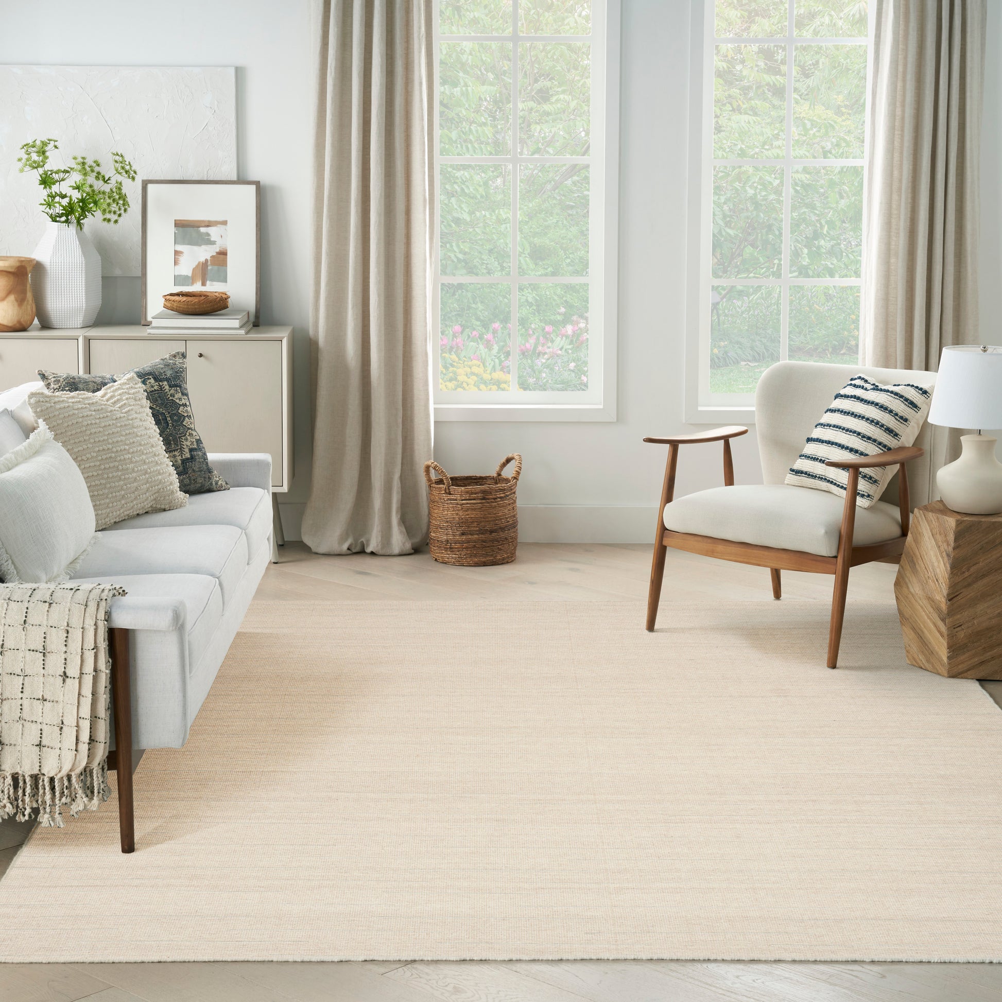 Bright living room with beige 8' x 10' rug, white sofa with textured pillows, mid-century armchair, woven basket, large windows.