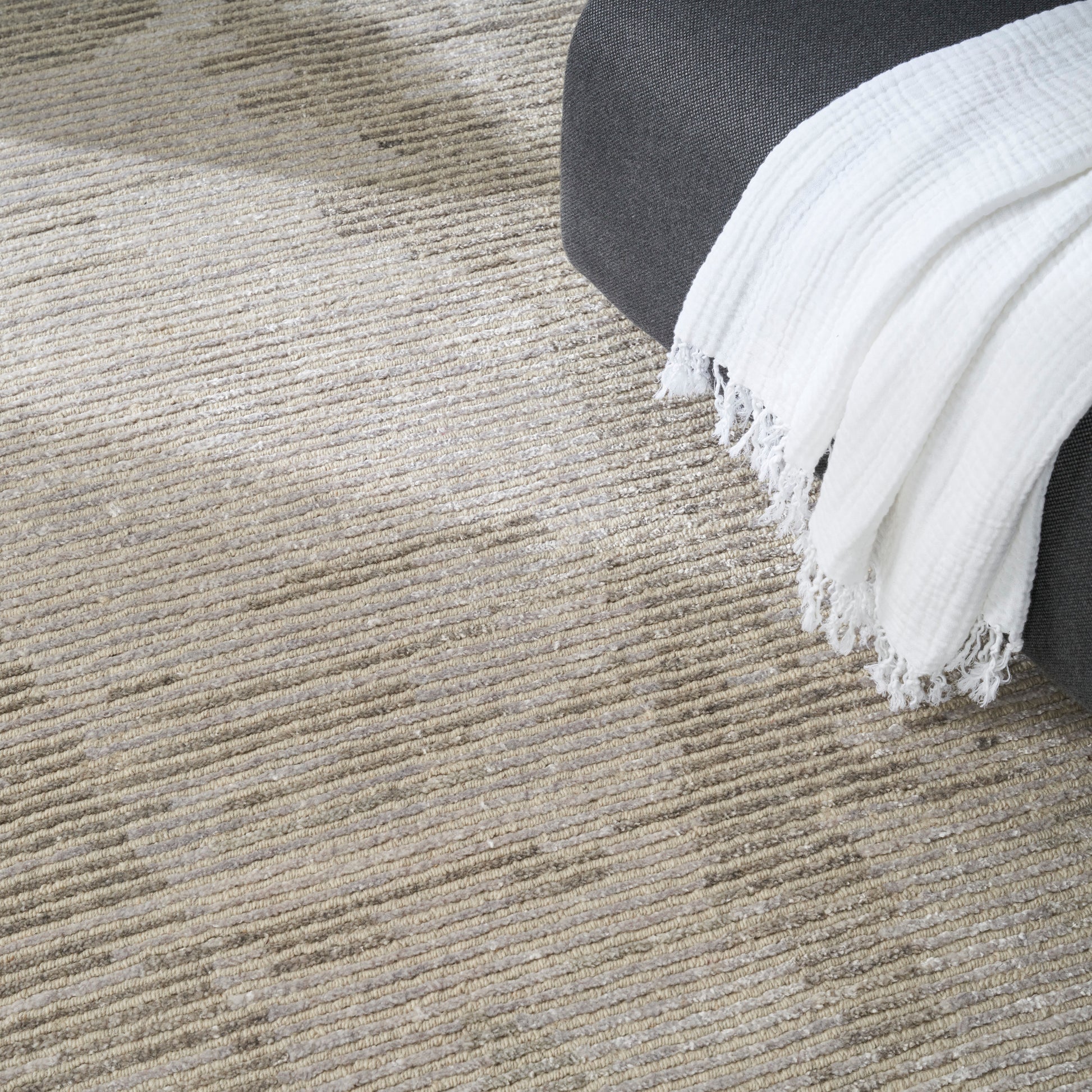 Beige textured rug with subtle horizontal stripes, gray sofa edge and white fringed throw blanket.