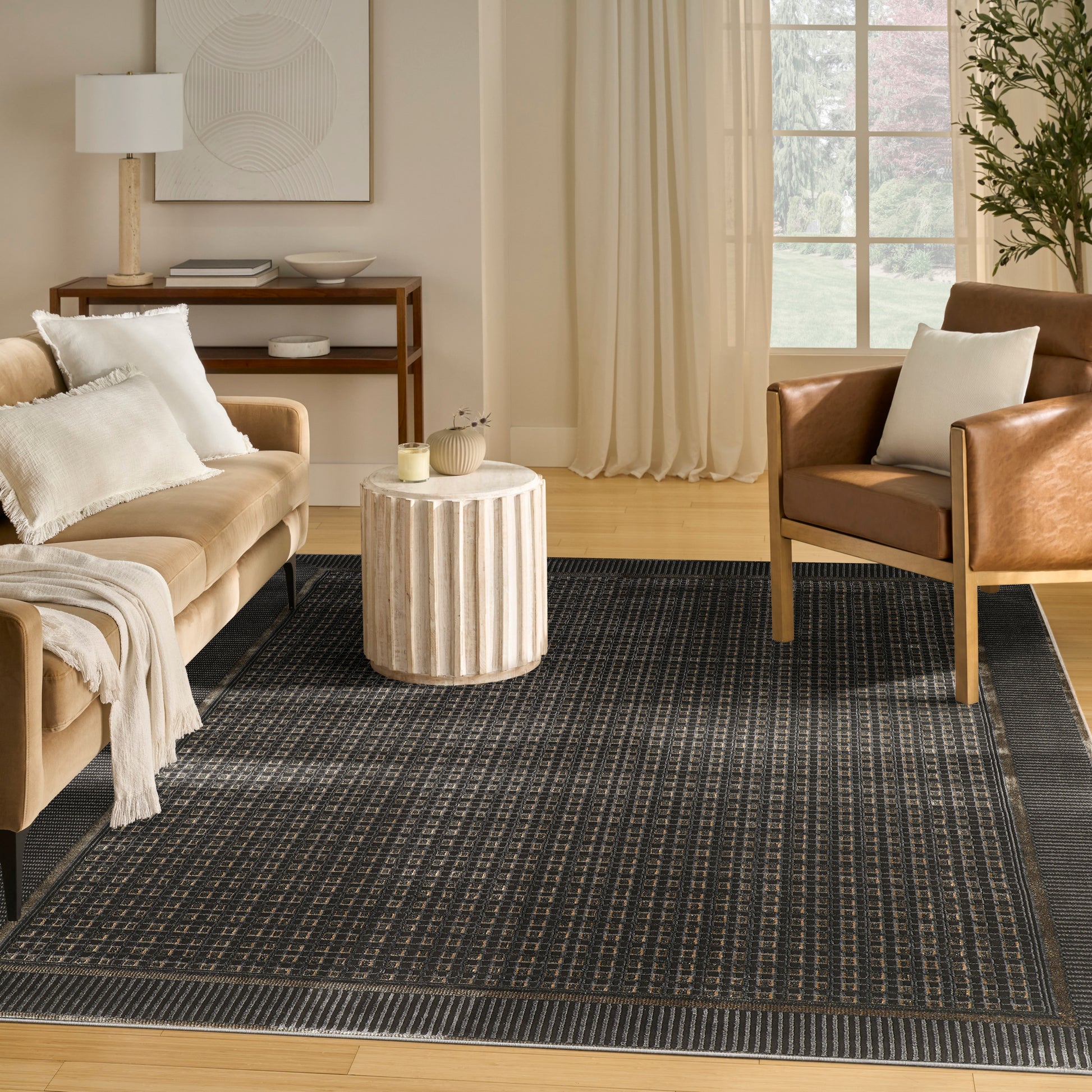 Cozy living room with tan sofa, brown leather armchair, round beige side table, and dark patterned rug.