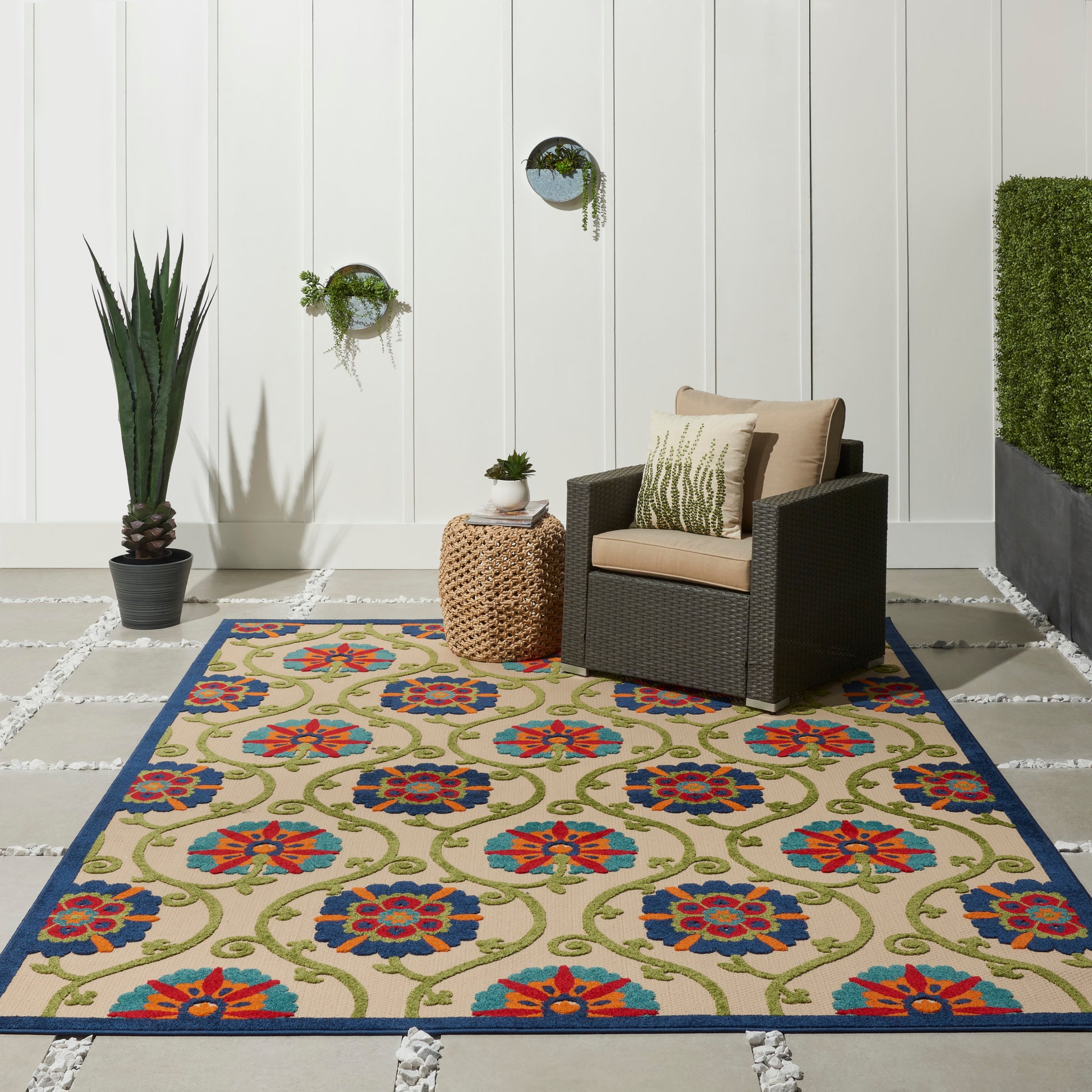 Outdoor patio with an 8' x 11' blue-bordered floral rug, dark wicker chair with tan cushions, round woven side table, and a tall plant.