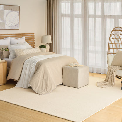 Neutral beige bedroom with wooden headboard, layered bed linens, cream rug, wicker chair, large sheer-curtained windows.