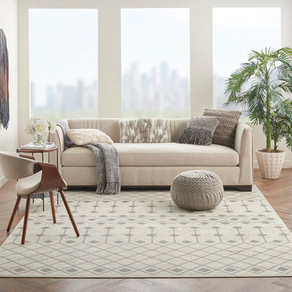 Beige fabric sofa with gray throws on a geometric gray rug, knitted pouf, wooden chair, and plant in living room.