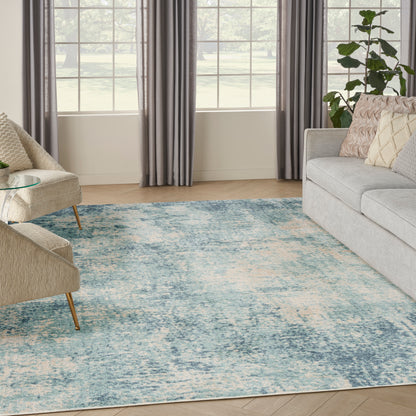 Bright living room with blue abstract area rug, beige accent chairs, gray sofa, glass coffee table, and large windows.