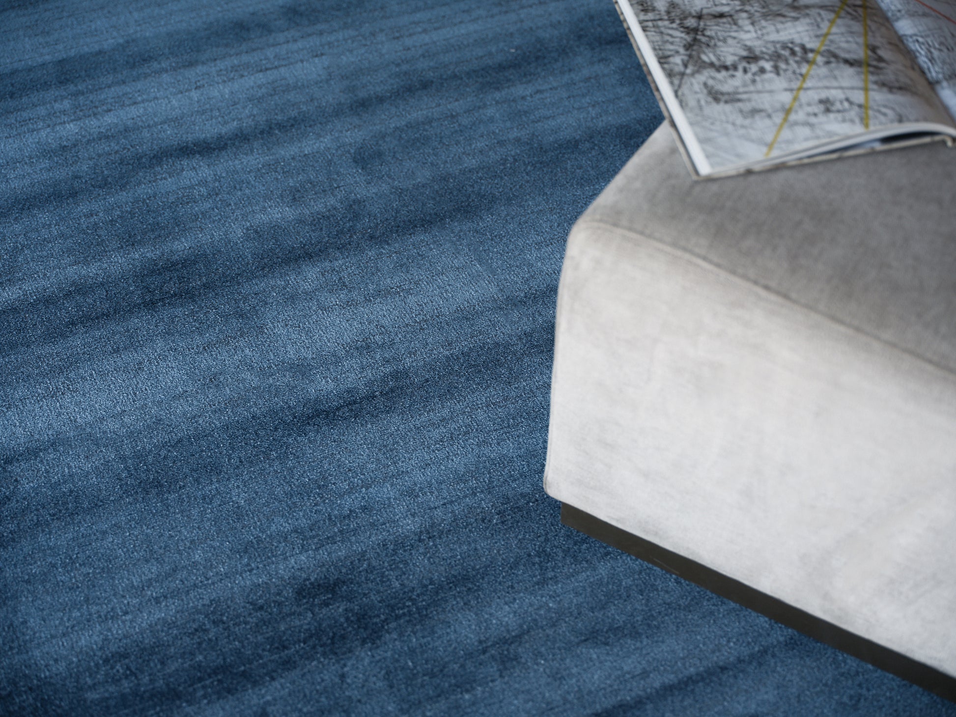 Blue textured rug beside a light gray upholstered ottoman with a magazine resting on its edge.