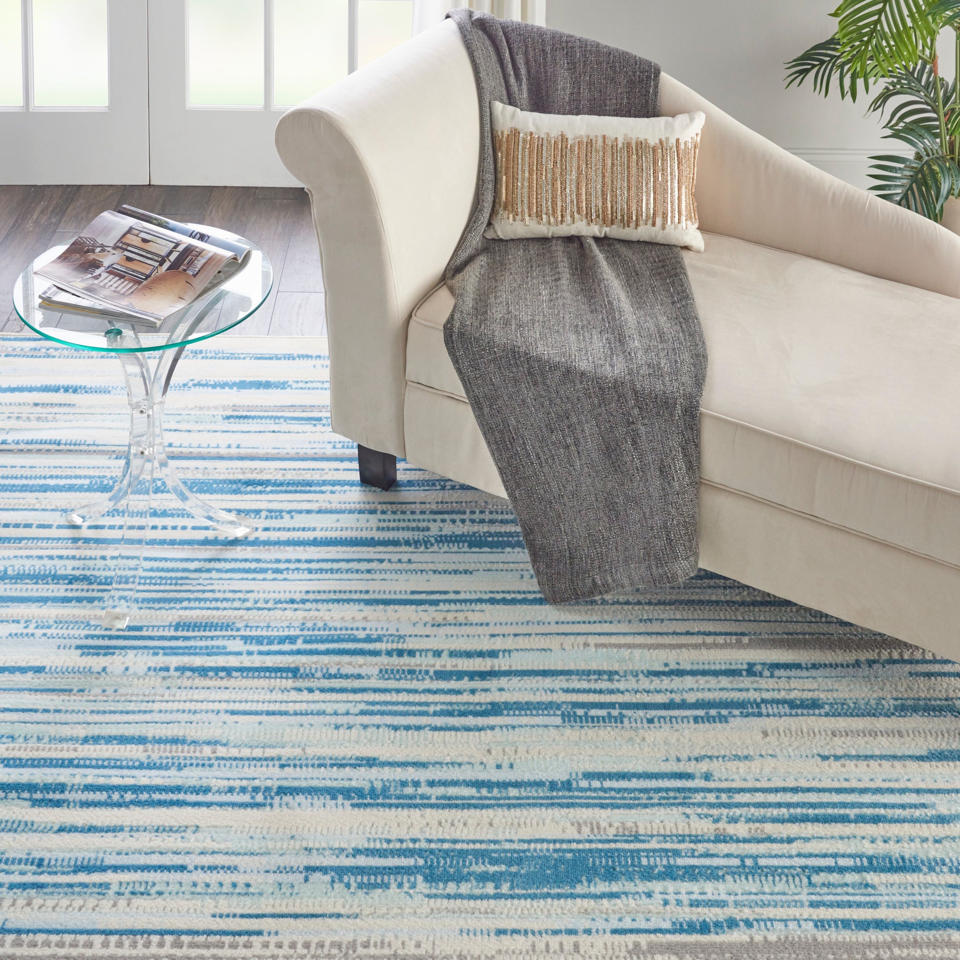 Beige sofa with gray throw and fringed pillow, round glass side table, blue striped rug in bright living room.