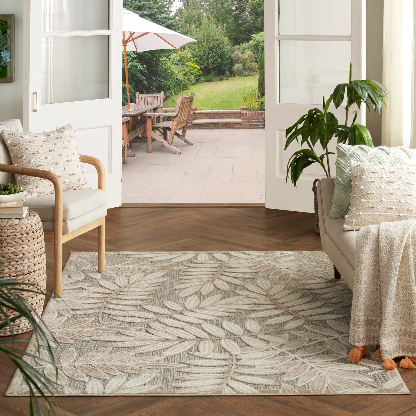 Neutral living room with a leafy gray-beige area rug, cream sofa, wooden chairs, and potted plants, open doors to patio