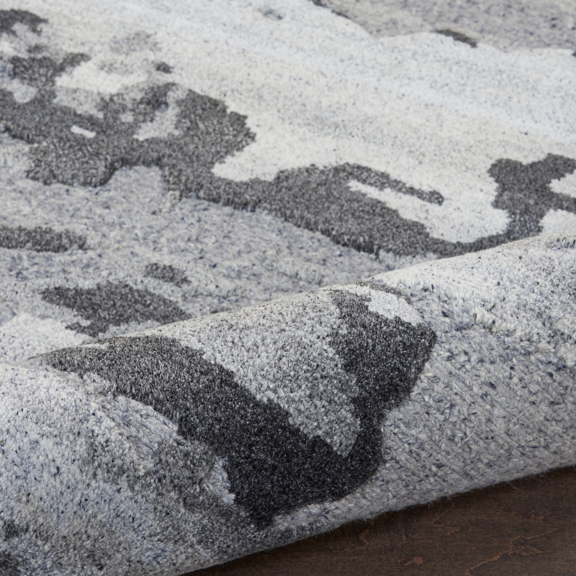 Shaggy area rug with abstract gray and charcoal patches; plush pile, folded corner on dark wood floor.