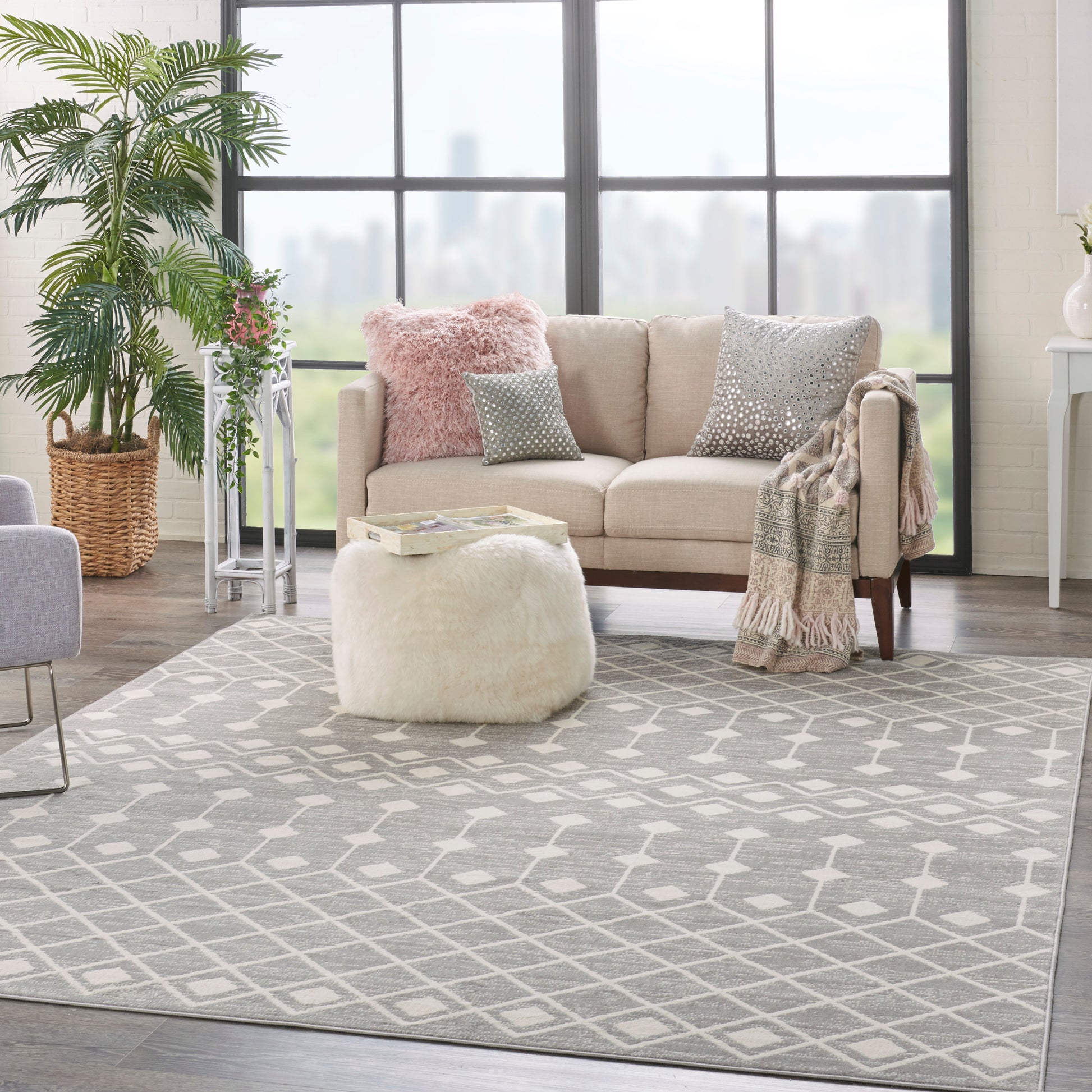 Bright living room with beige sofa, pink fuzzy pillow, gray geometric rug, round cream ottoman, throw blanket, and plants.