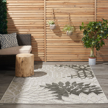 Outdoor patio with wicker sofa, beige cushions, round wood stump table, leaf-pattern rug, hanging planters, potted tree.