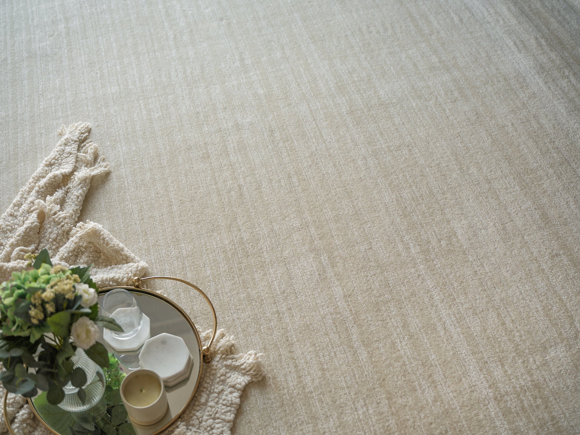 Beige textured rug with cream fringe; round gold-trim mirror tray holding candles and a floral arrangement.