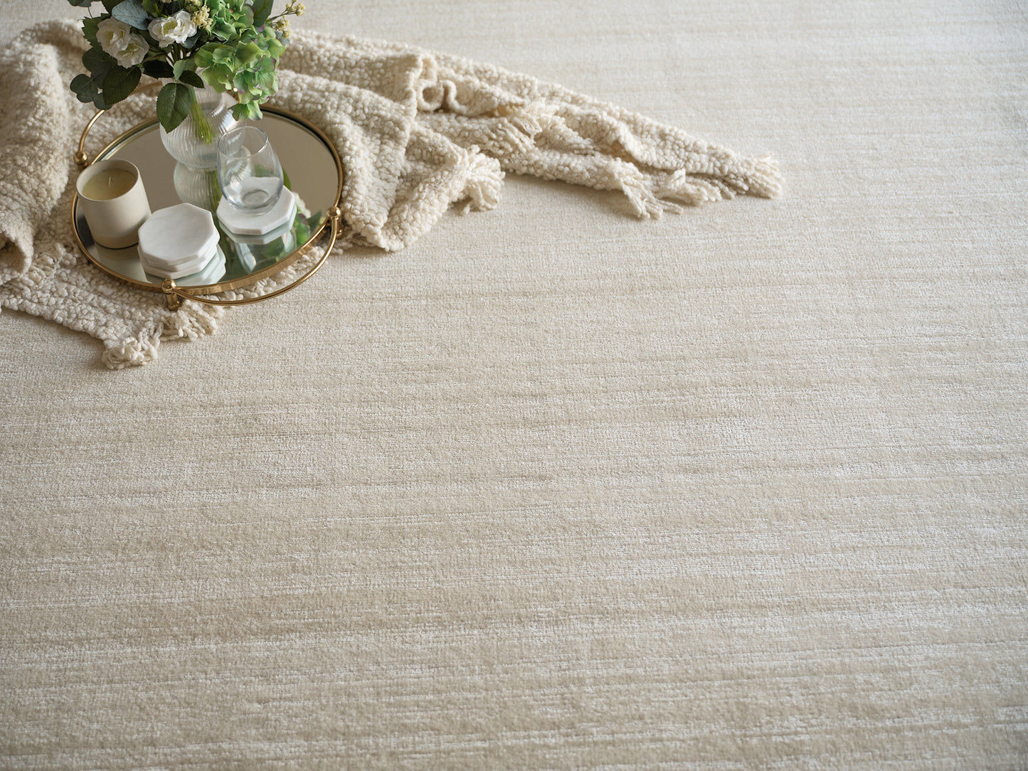 Beige textured rug with round gold-trim mirrored tray, candles, marble candle holder, glass vase, and cream fringe blanket.