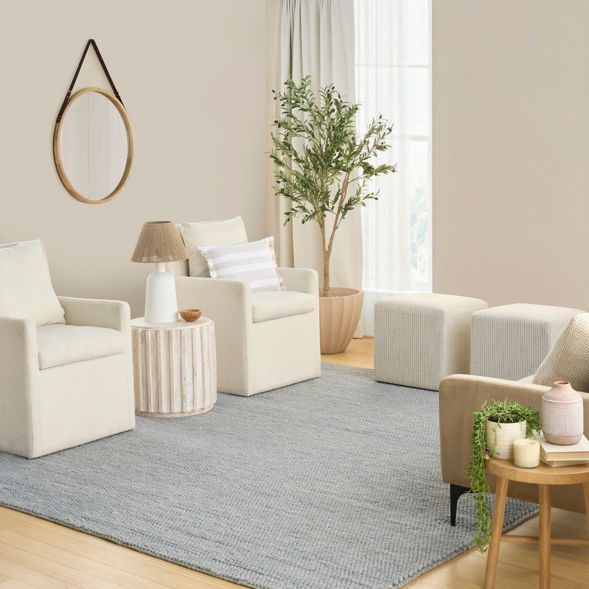 Gray rug in a neutral living room with cream armchairs, a round mirror, woven side tables, a potted plant and light curtains.