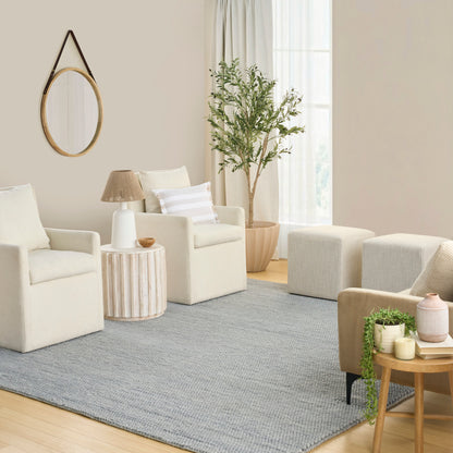 Gray rug in a neutral living room with cream armchairs, a round mirror, woven side tables, a potted plant and light curtains.