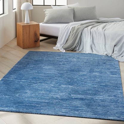 Blue textured rectangular area rug in a modern bedroom with gray bedding, light wood nightstand & white lamp.