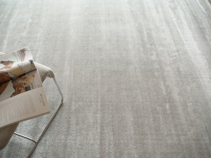 Light gray textured area rug with a soft woven look; corner shows a beige chair with metal legs & an open magazine.
