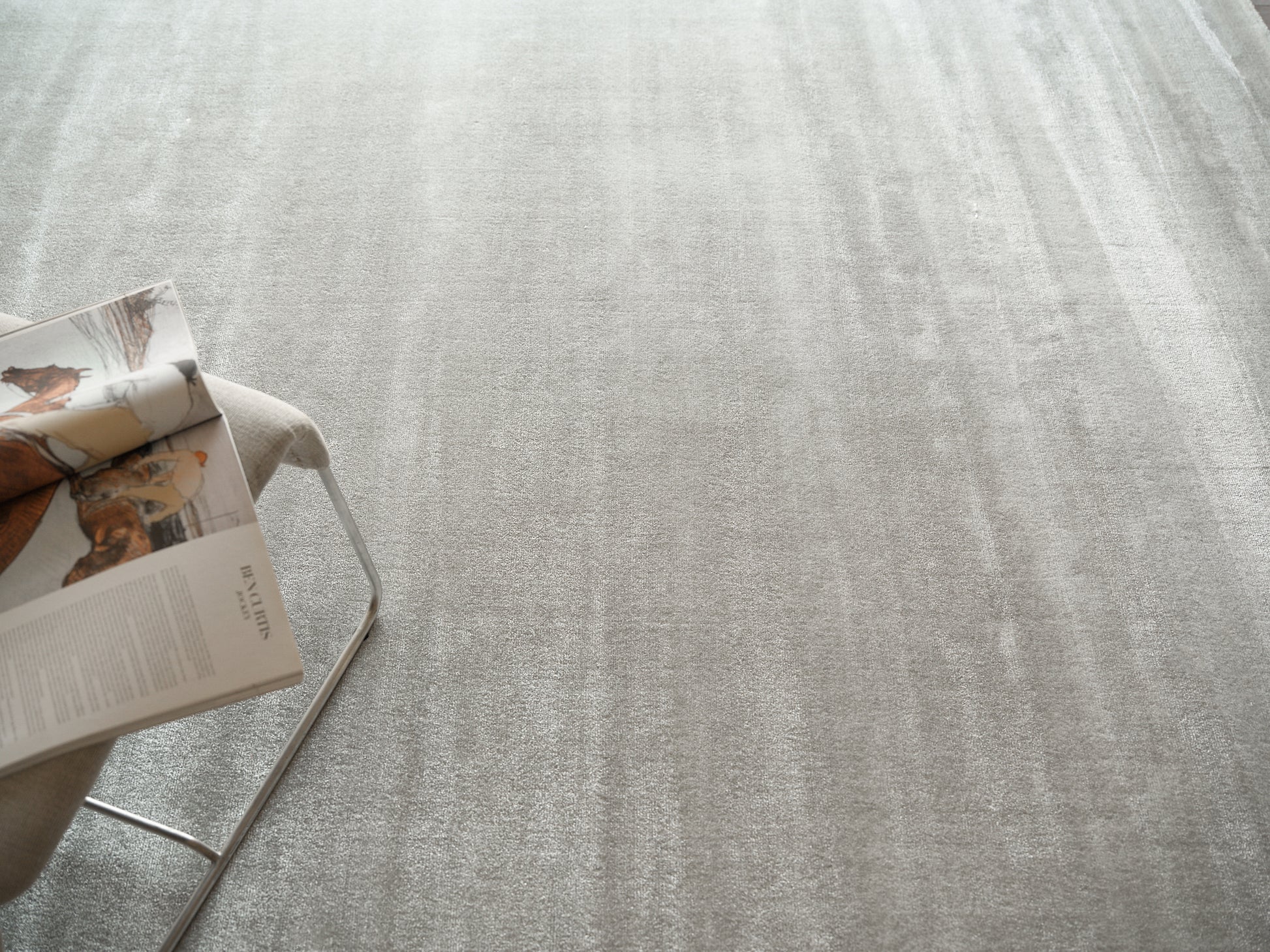 Light gray textured rug with soft pile; beige fabric on a minimalist metal-framed chair with an open magazine.