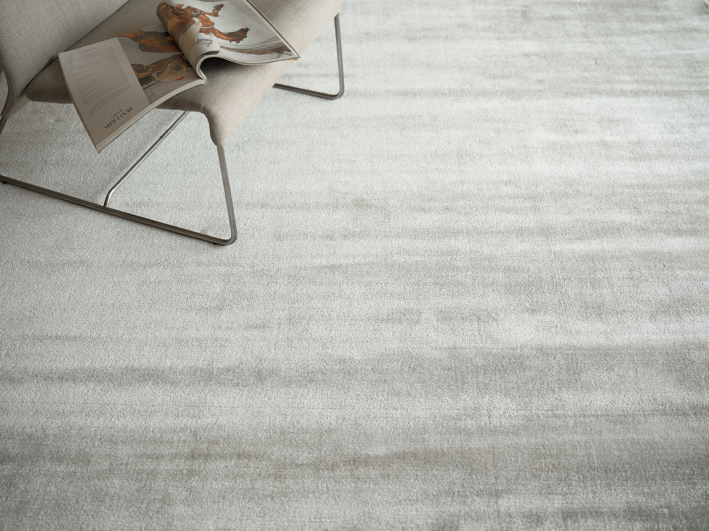 Light gray textured area rug with a modern metal-framed lounge chair and an open magazine resting on the chair.