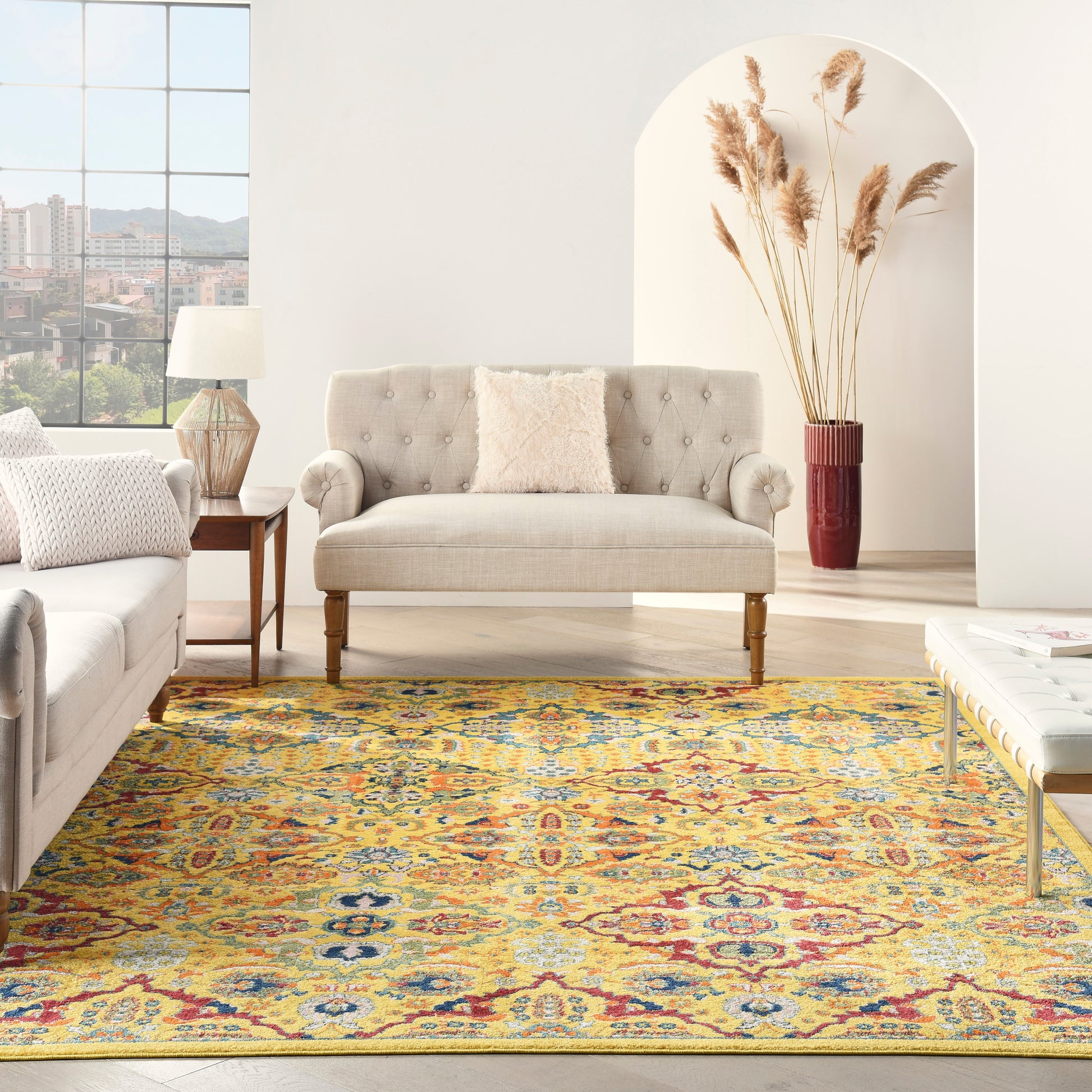 Living room with a beige tufted sofa on wooden legs, a light gray sofa, side-table lamp, and a colorful yellow-blue-red Persian rug, pampas grass vase by an arch alcove.