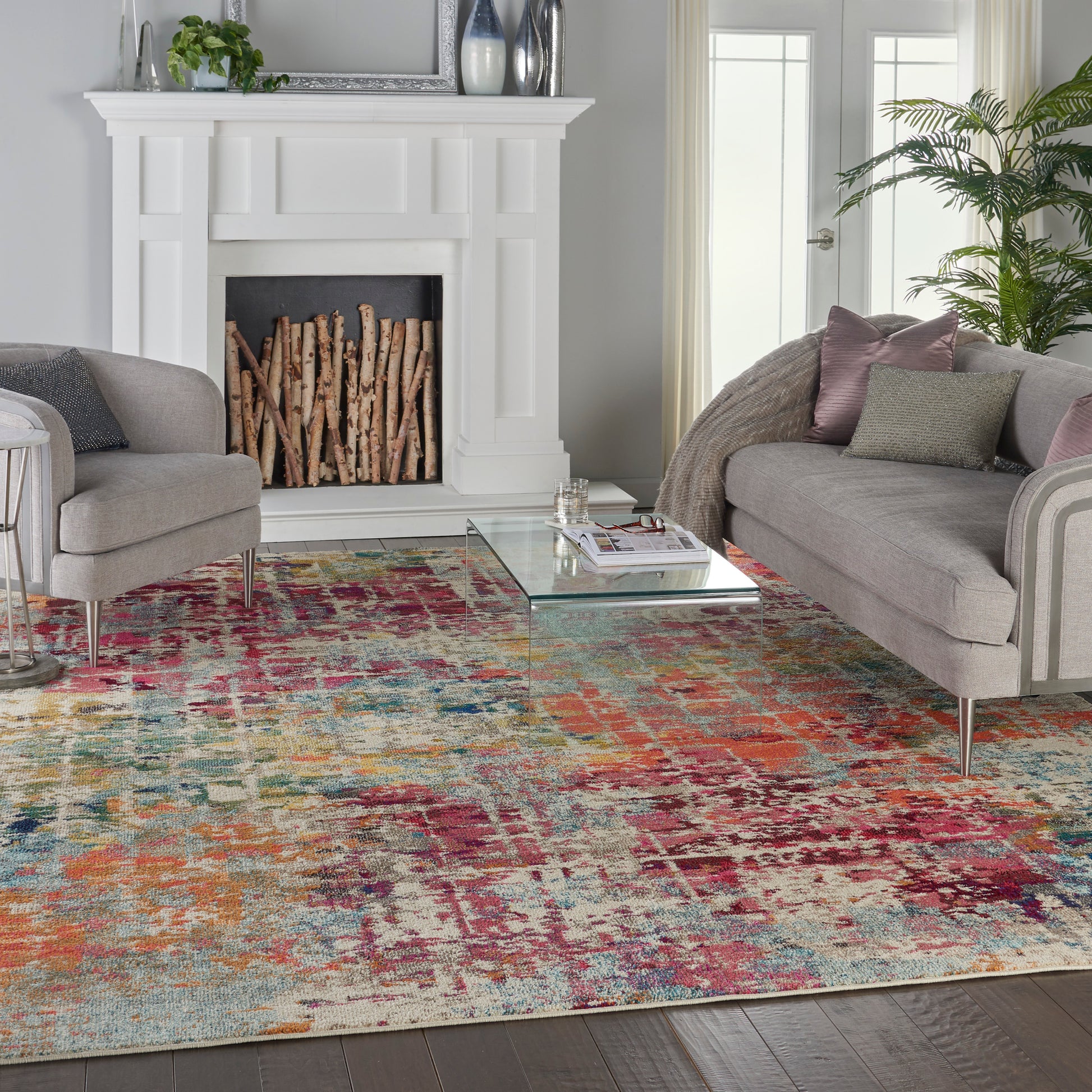 Colorful abstract area rug in pinks, blues, oranges on a dark wood floor; glass coffee table, gray sofas, white fireplace.