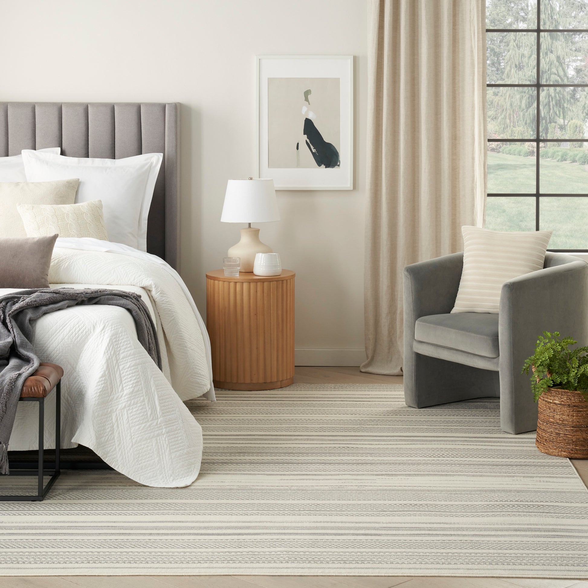 Neutral-toned rug in a bedroom scene with a tufted gray bed, white bedding, beige pillows; round wooden nightstand with a lamp, and a gray armchair.