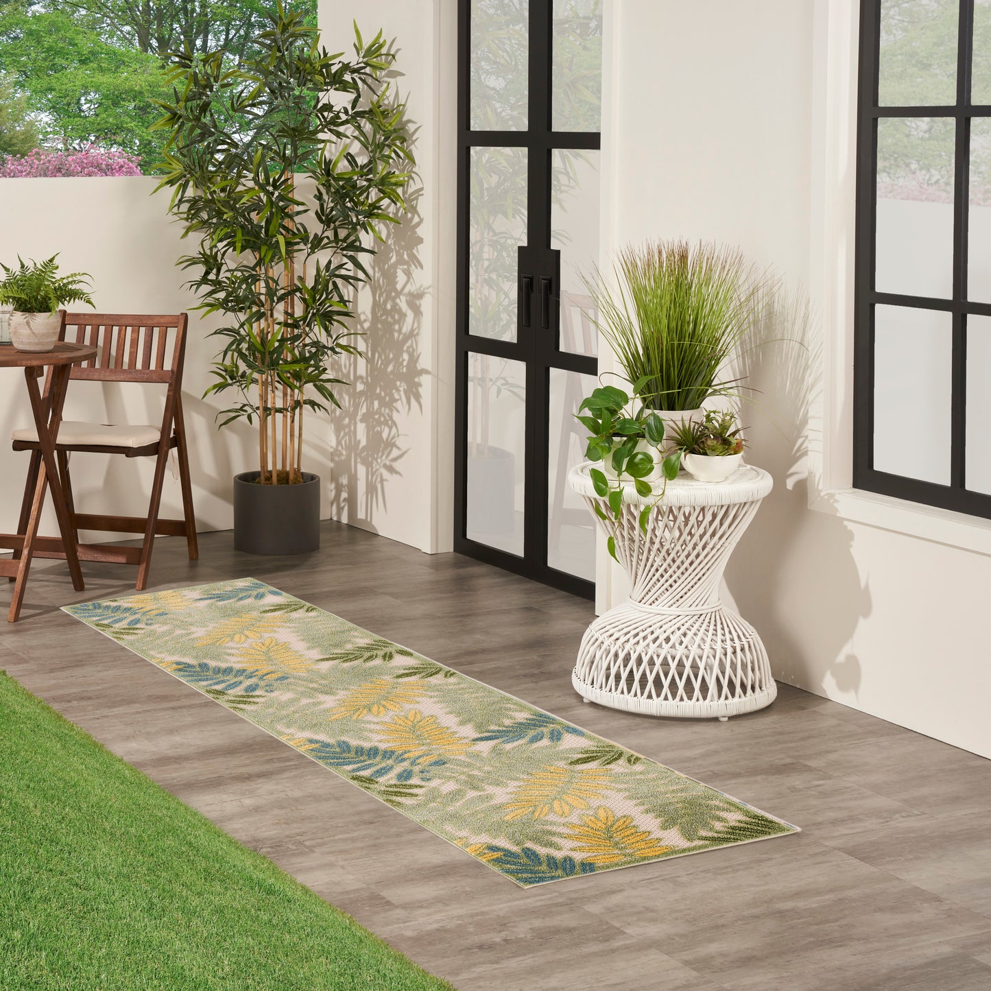 Long botanical-pattern runner rug with green, yellow, teal leaves on gray background beside indoor plants and glass doors.
