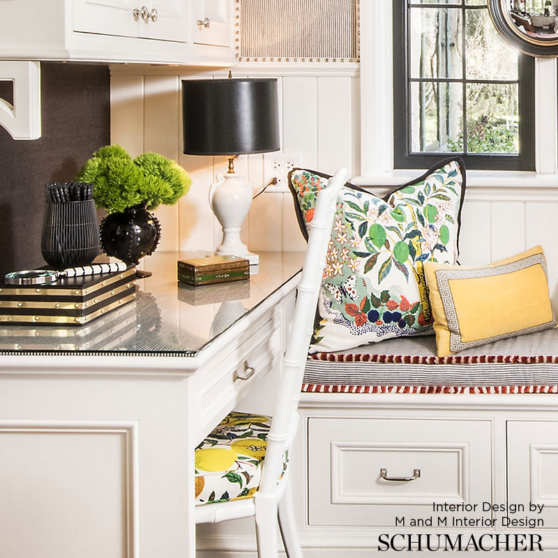 White kitchen desk nook with glass top, black lamp, green plants, stacked books, white chair, and floral and yellow pillows.